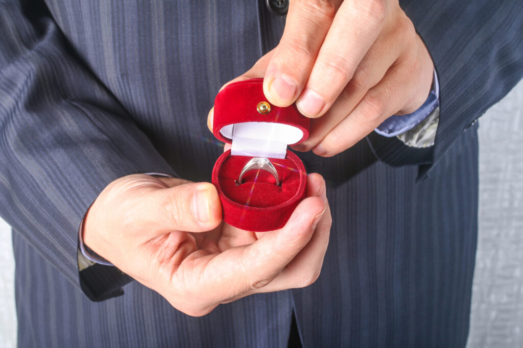 Man in a suit holding an open red velvet ring box with a diamond engagement ring inside, symbolizing smart engagement ring shopping and budgeting.
