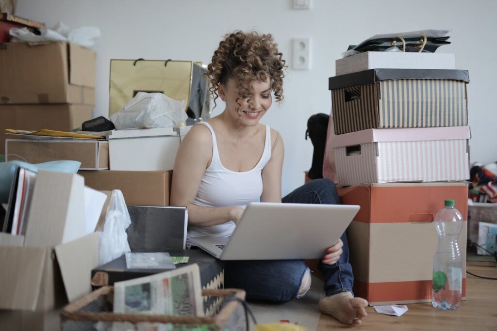 A woman sitting on the floor surrounded by boxes, looking at a laptop.
