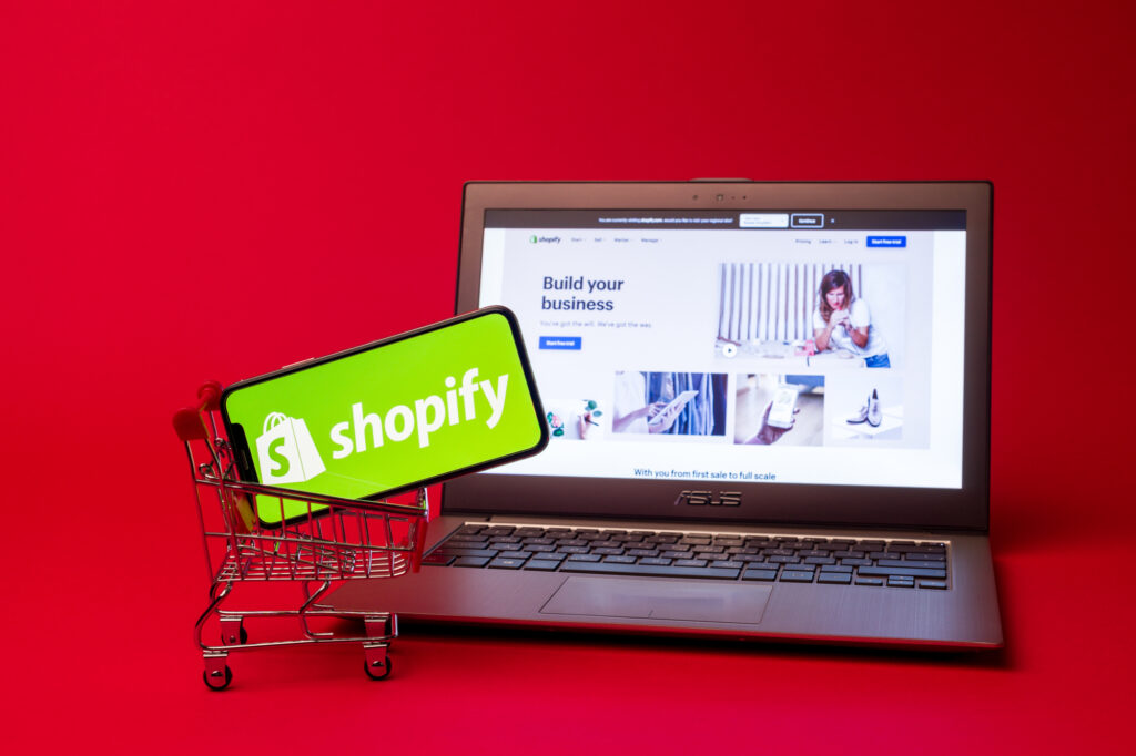 Tula, Russia - AUGUST 19, 2019: Shopify web page displayed on a modern laptop on red background. Shopify logo on iphone X in shopping cart