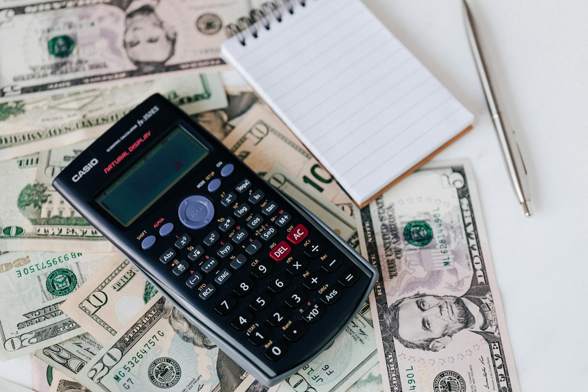 A notepad, pen, calculator, and dollar bills. 