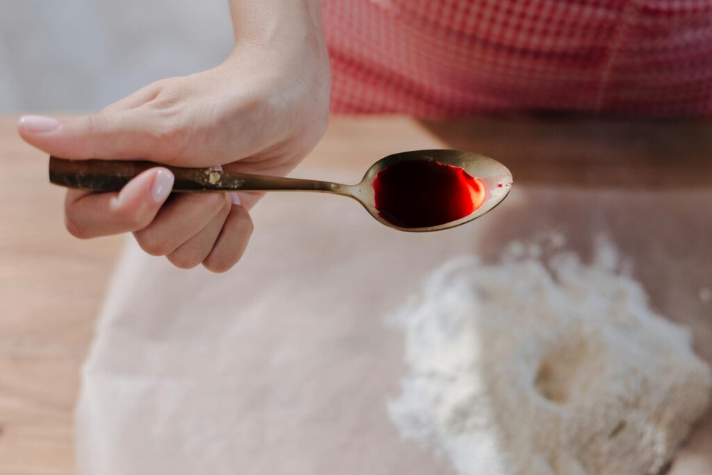 A hand holding a spoon filled with red food dye over a surface with flour.