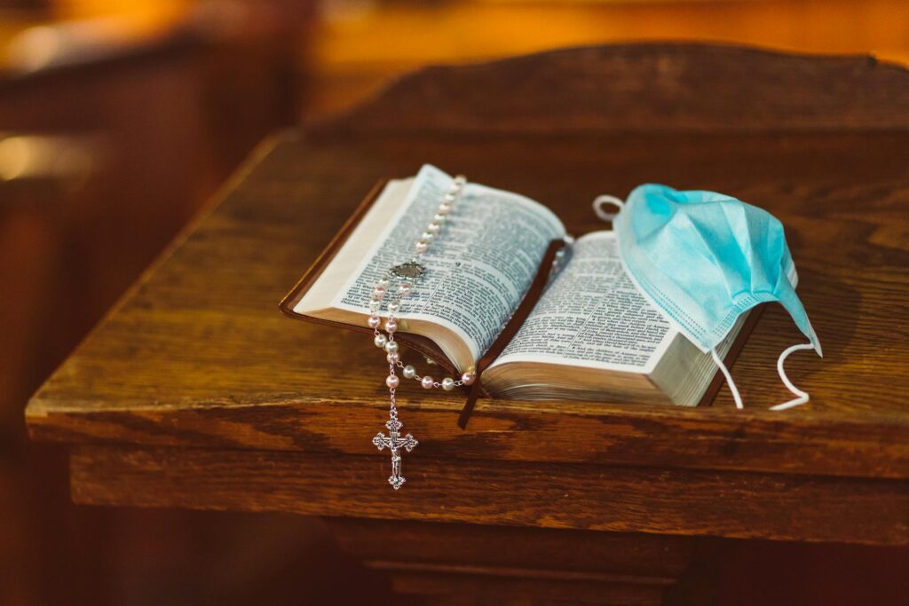 An open Bible on a wooden pulpit with a rosary and a blue medical face mask resting on its pages, symbolizing the intersection of faith and healthcare.