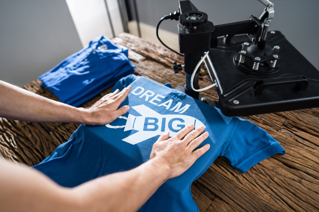 Hands pressing a "Dream Big" design onto a blue t-shirt using a heat press machine on a wooden table.