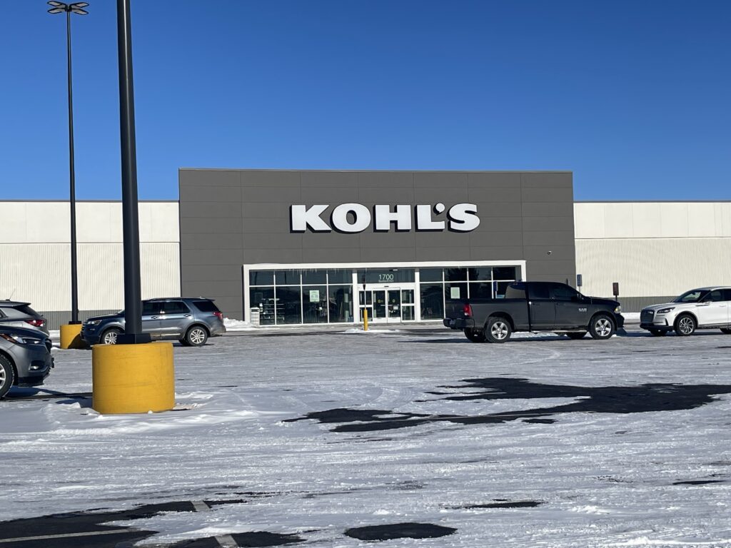 A Kohl's department store building with a dark gray facade and large white signage, surrounded by a snowy parking lot with several parked vehicles.