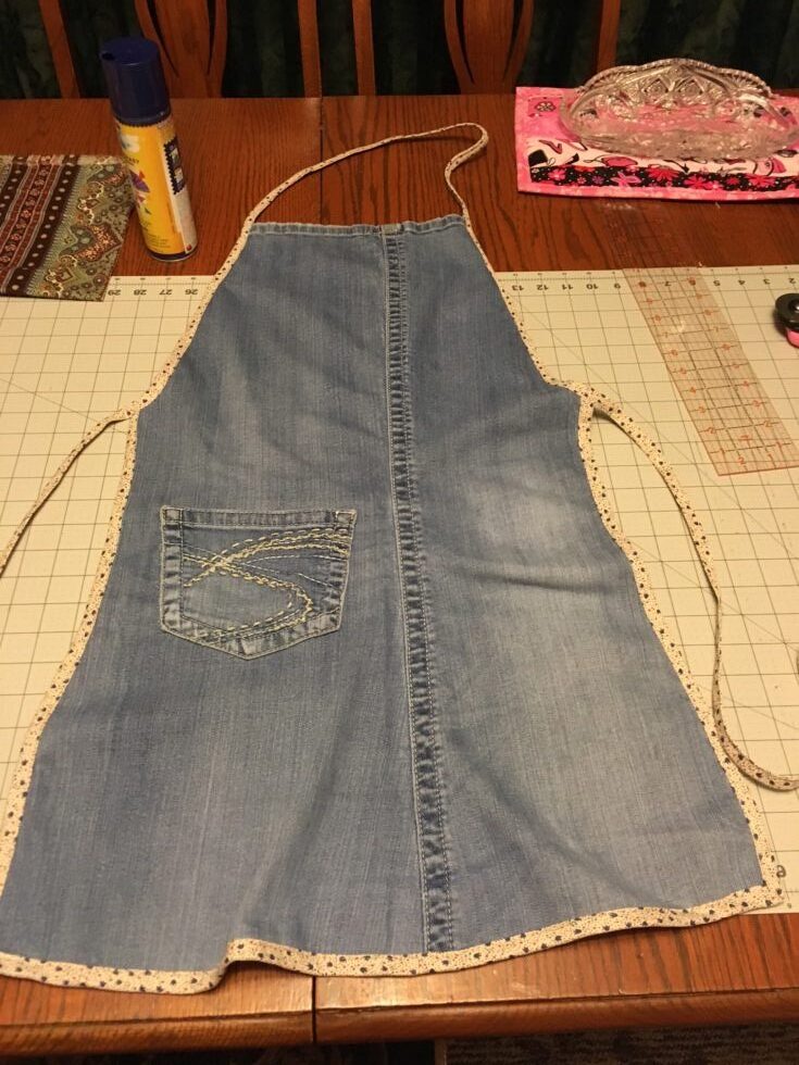 A handmade apron crafted from repurposed denim jeans, featuring a decorative pocket and fabric trim, laid out on a crafting table.