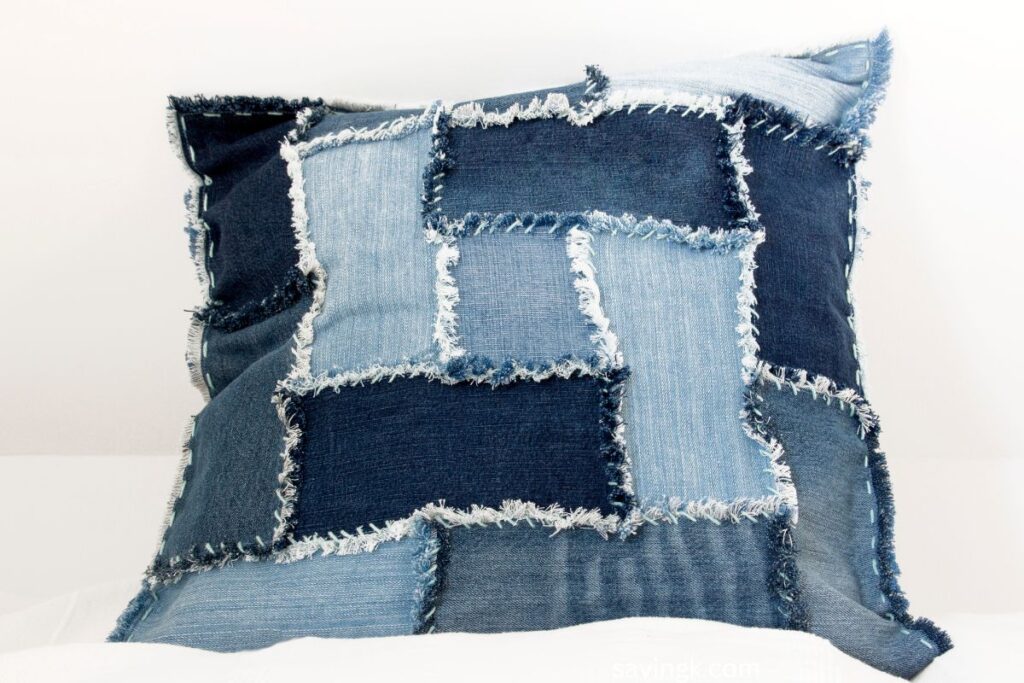 A handmade patchwork pillow made from repurposed denim fabric, featuring frayed edges and varying shades of blue denim.