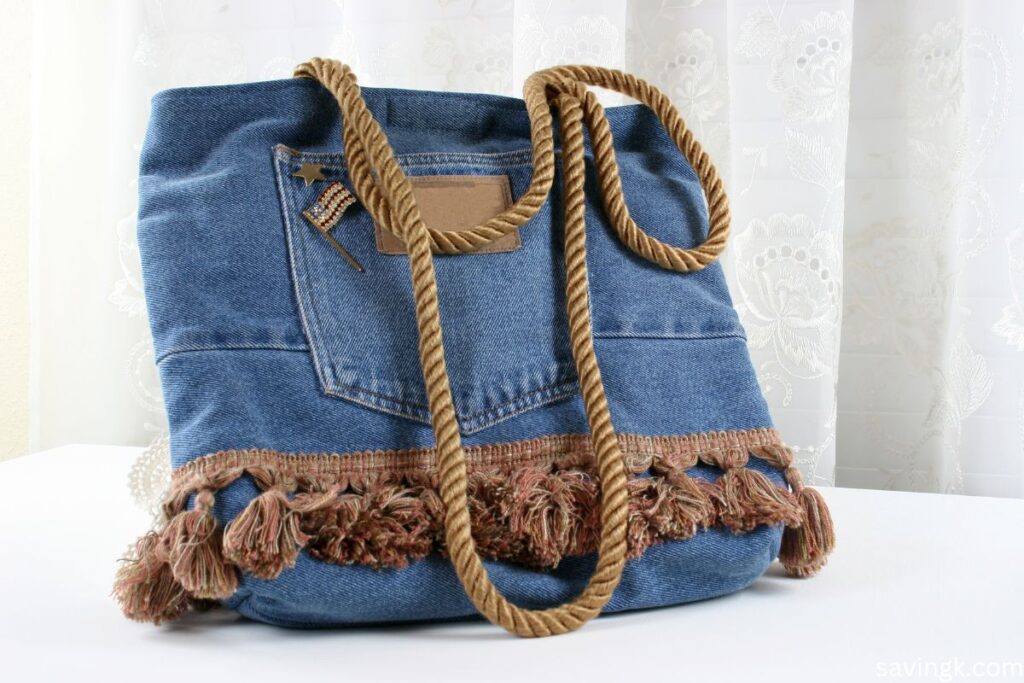 A handmade denim purse crafted from repurposed jeans, featuring a front pocket, decorative fringe, and rope handles.