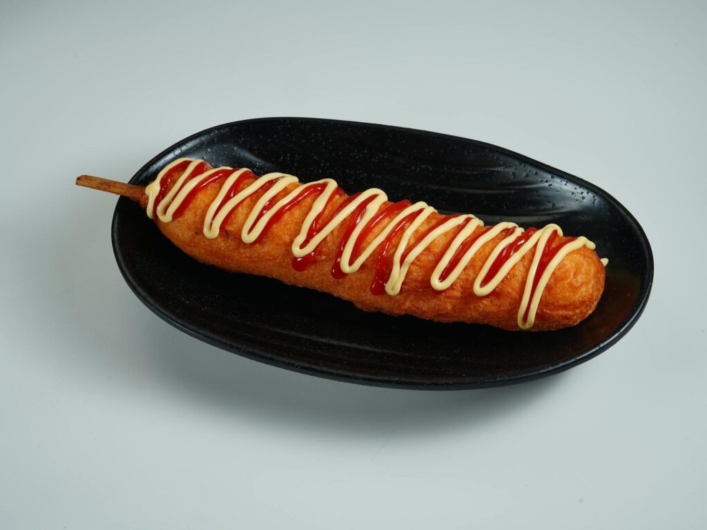 A crispy golden corndog drizzled with ketchup and mayonnaise, served on a black plate against a white background.