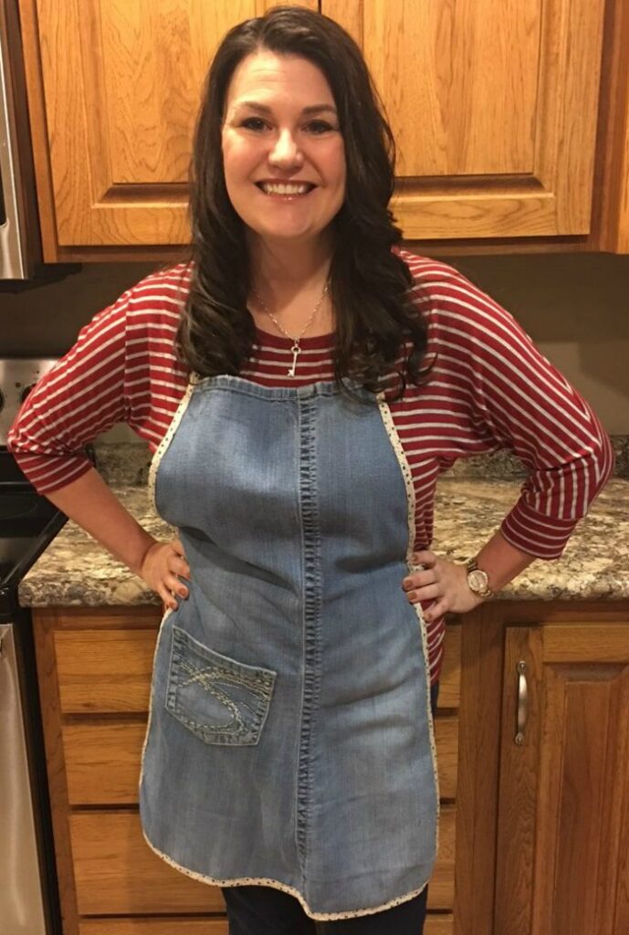 A smiling woman wearing a handmade denim apron repurposed from old jeans, standing in a kitchen with wooden cabinets and a granite countertop.