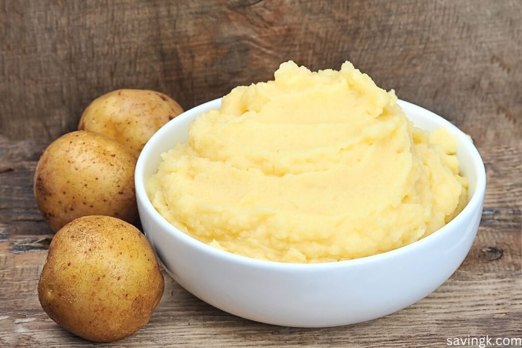 A bowl of creamy mashed potatoes in a white dish, surrounded by raw potatoes on a rustic wooden surface.