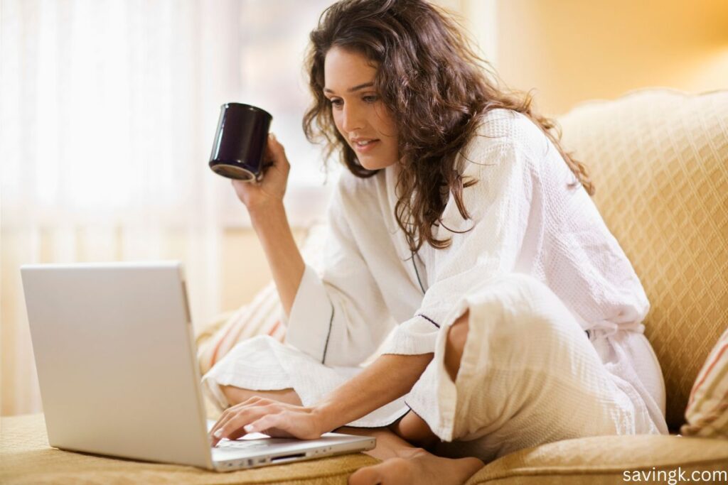 Woman in a white robe sitting on a couch, drinking coffee while browsing on a laptop.