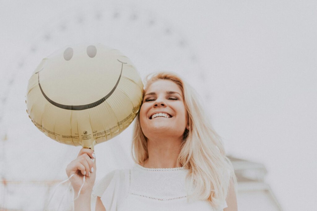 A smiling blonde woman holding a yellow smiley face balloon near her head, with a blurred Ferris wheel in the background.