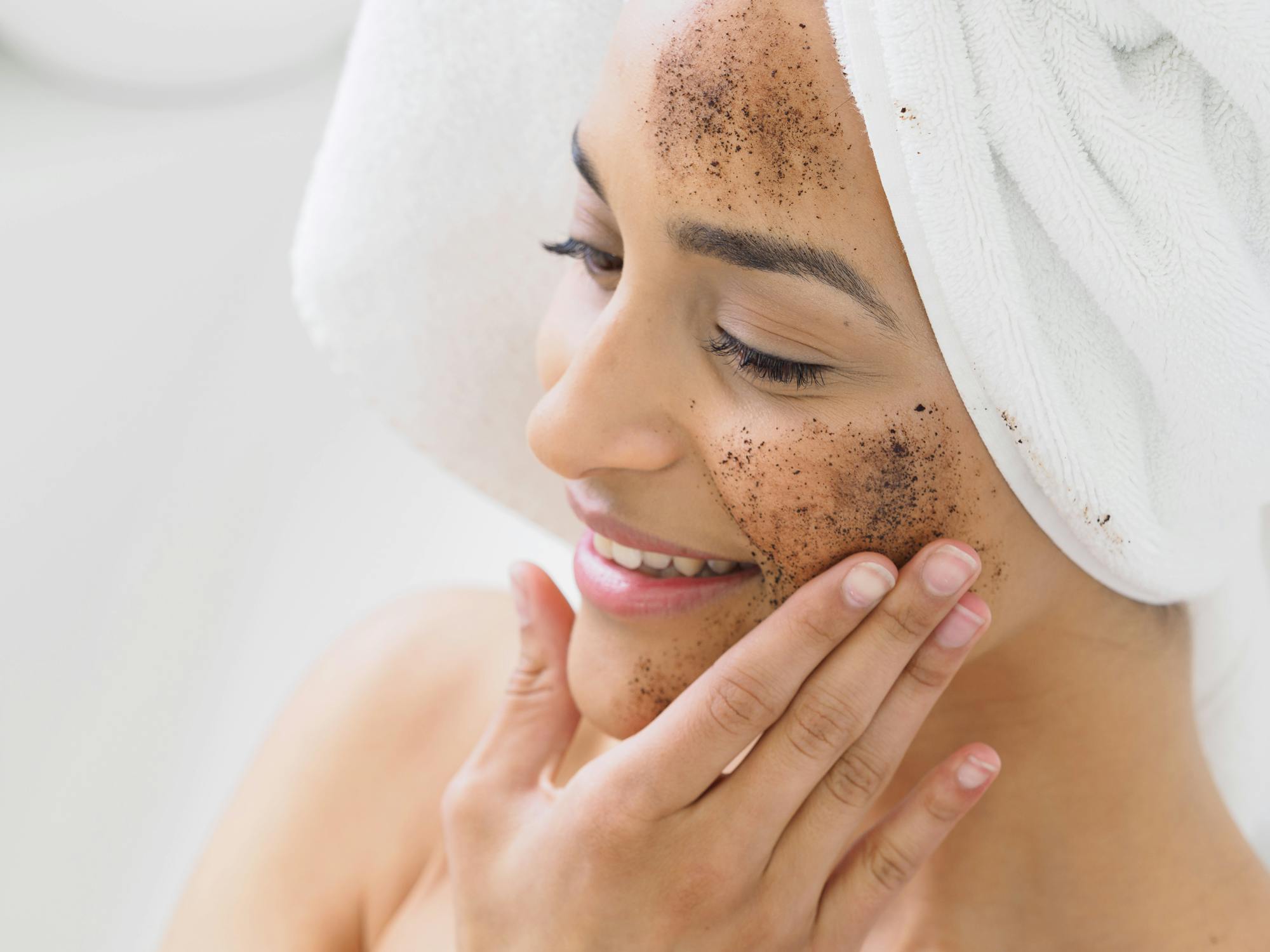 A smiling woman with a towel wrapped around her head applies a coffee scrub to her face for exfoliation.