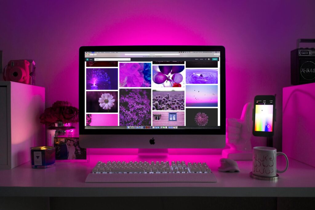 A stylish workspace featuring an Apple iMac displaying a gallery of purple-themed images, illuminated by vibrant pink LED lighting. A white keyboard and mouse sit on the desk, along with a coffee mug, a smartphone on a stand, and various decorative elements.