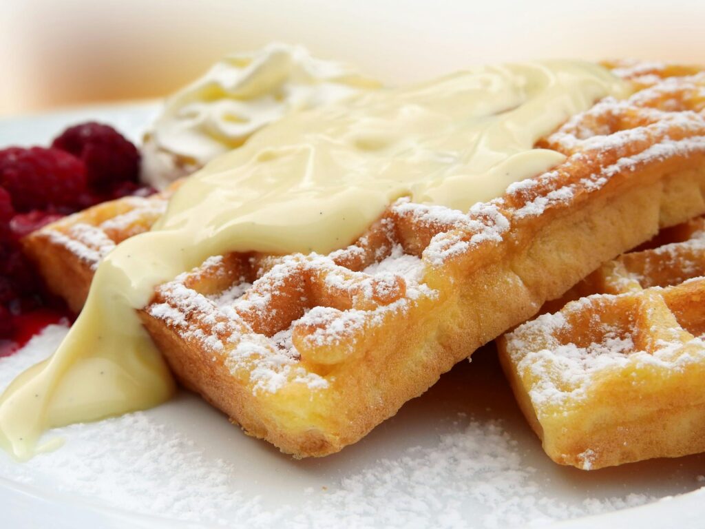 A close-up of golden, crispy waffles dusted with powdered sugar, topped with creamy vanilla sauce, and served with fresh raspberries and whipped cream.