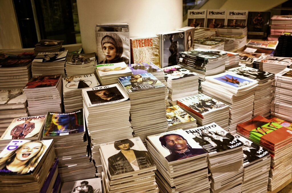 Stacks of various fashion and lifestyle magazines piled high on a table, featuring colorful and stylish covers with models and celebrities.