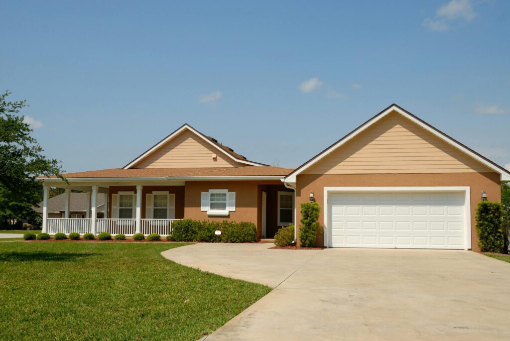 A single-story suburban house with a beige and brown exterior, a white garage door, and a wraparound porch, surrounded by a green lawn under a clear blue sky.