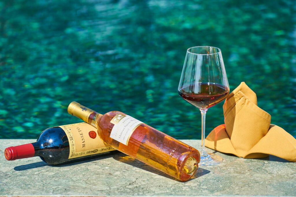 Two bottles of wine, one red and one white, resting on a stone surface near a swimming pool. A glass of red wine and a neatly folded yellow napkin sit beside them, reflecting a luxurious and relaxing outdoor setting.