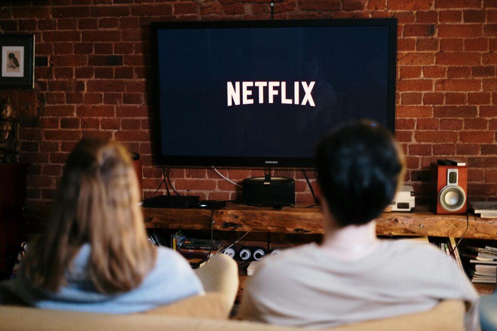 Two people sitting on a couch watching Netflix on a TV mounted on a brick wall, with a wooden entertainment center holding various electronic devices and speakers.