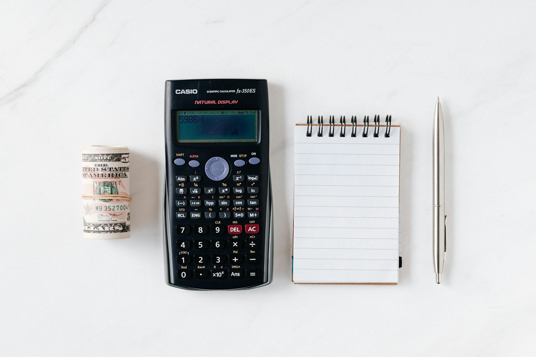 Rolled bills, calculator, notepad and a pen on a white surface