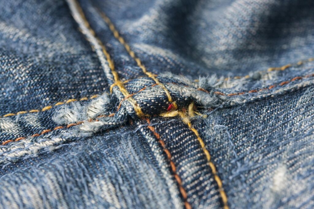 A close-up view of worn and frayed denim fabric with detailed stitching and distressed seams.