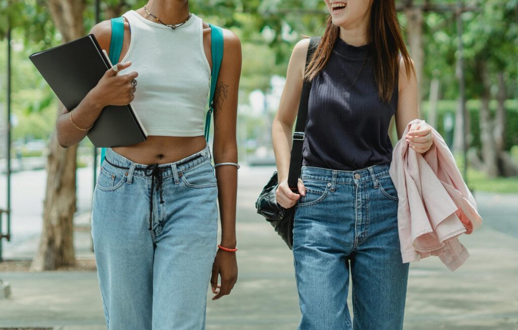 Two young women in casual outfits walking outdoors on a college campus, smiling and chatting while carrying a laptop and a jacket.