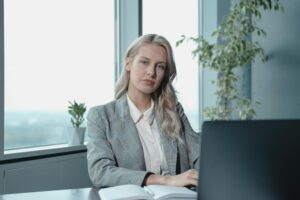 A confident businesswoman with long blonde hair, wearing a gray plaid blazer and white blouse, sits at a desk with a laptop and an open notebook in a modern office with large windows and greenery in the background.