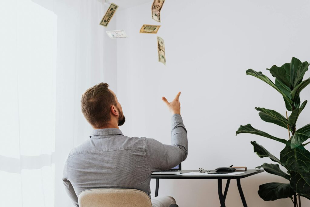 A man sitting at a desk throws money into the air, symbolizing wasteful spending or financial freedom.