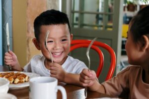 A young boy smiling while holding a fork and knife at the breakfast table, with waffles on his plate and a friend sitting across from him.