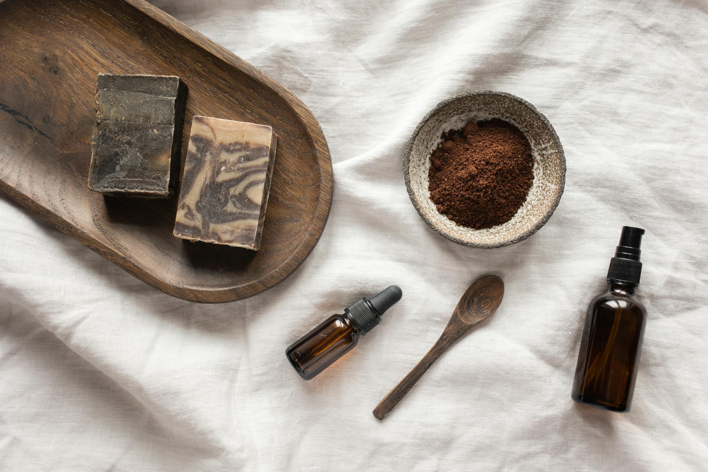 A flat lay of natural skincare products, including handmade coffee-infused soap, a bowl of ground coffee, essential oils, and a wooden spoon on a linen fabric background.