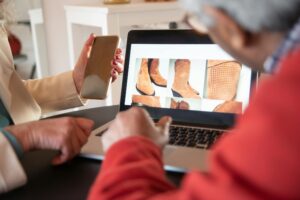 Two people browsing an online store on a laptop, looking at images of brown suede boots while discussing a potential purchase.