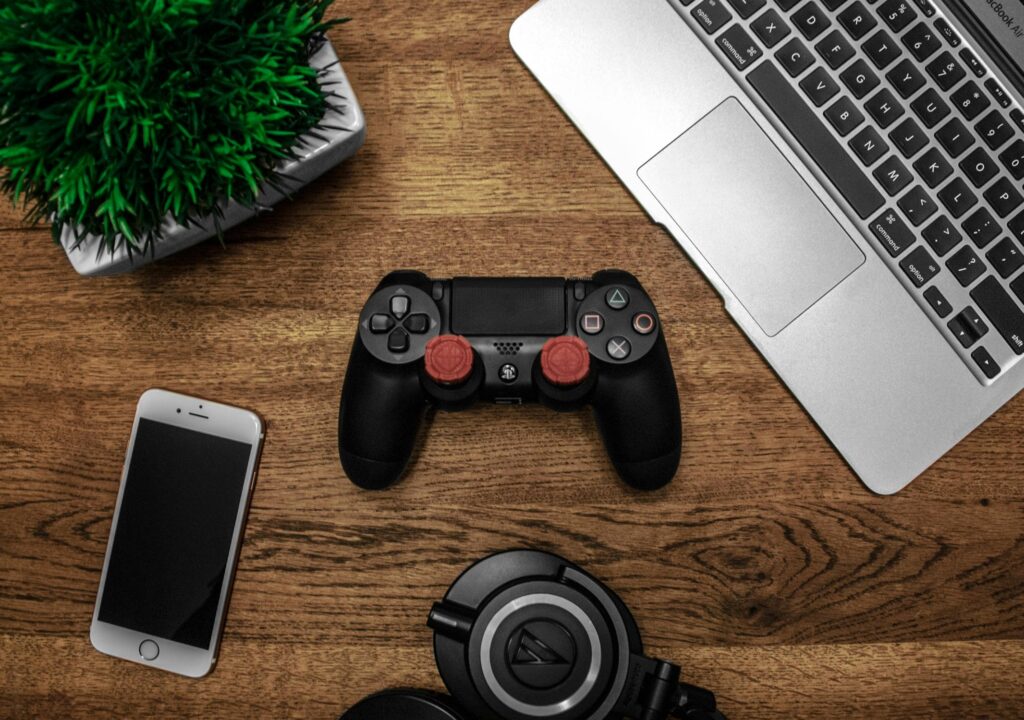 A gaming setup on a wooden desk featuring a PlayStation controller, a smartphone, a laptop, a pair of headphones, and a small green plant.