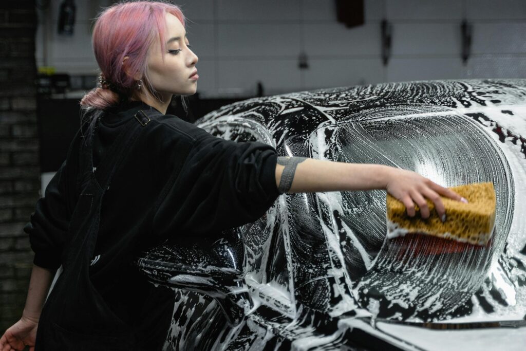 A young woman with pink hair washing a black car with a sponge and soap suds in a garage.