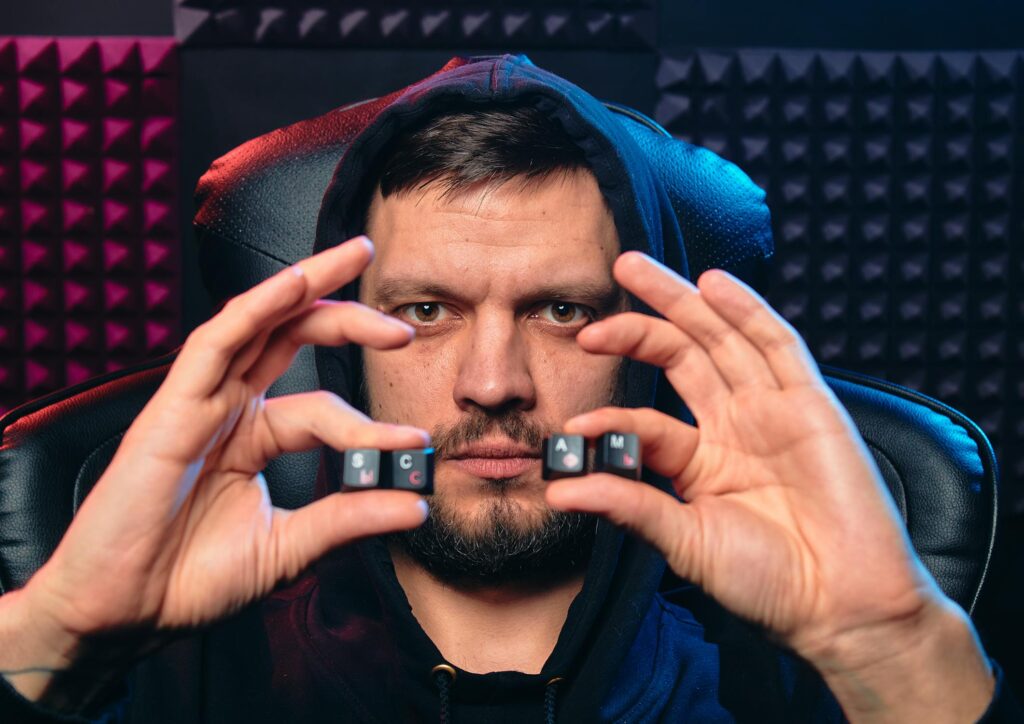 A man in a black hoodie sits in a gaming chair, holding keyboard keys that spell "SCAM" with an intense and serious expression, symbolizing cybercrime or financial fraud.