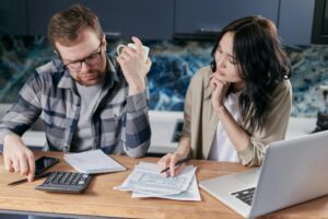 A stressed couple reviewing financial documents at their kitchen table, with a calculator, notebook, and laptop in front of them.