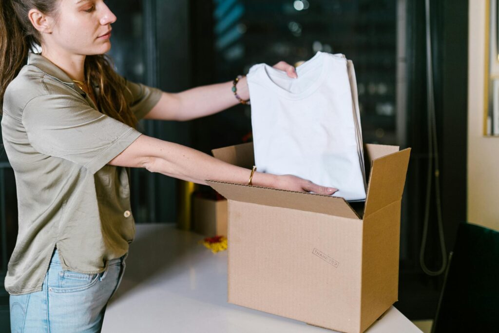 A woman packing a neatly folded white t-shirt into a cardboard box, preparing clothing for shipment.