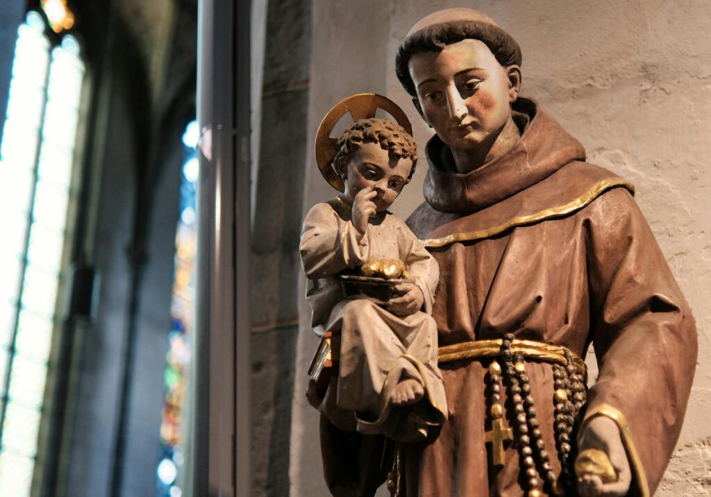 A statue of St. Anthony of Padua holding the child Jesus, adorned with a halo and wearing a brown Franciscan robe, inside a church with stained glass windows in the background.