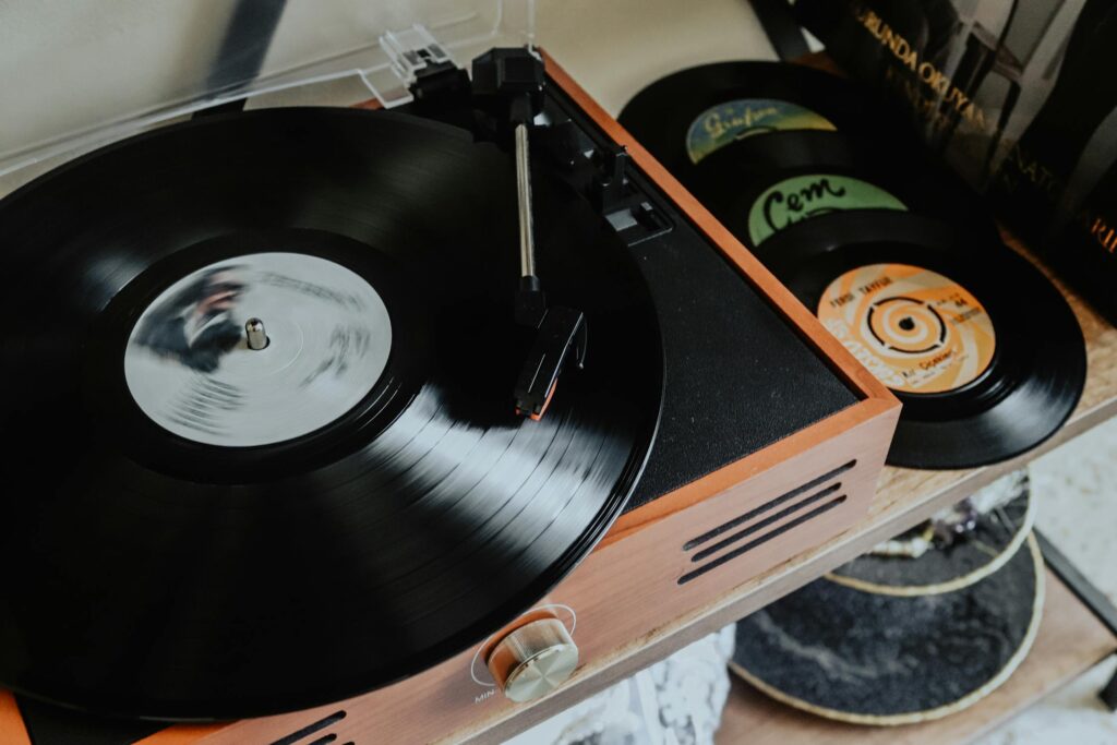 A vintage wooden record player spinning a black vinyl record, with several smaller vinyl records stacked beside it on a wooden shelf.