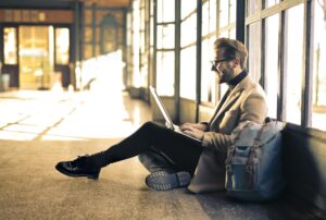 A stylish man with glasses, a beard, and a tan coat sits on the floor near large windows, working on his laptop with a backpack beside him.