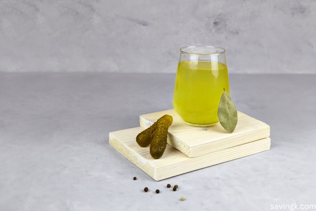 A glass of pickle juice on a wooden board, accompanied by pickles, a bay leaf, and scattered peppercorns.