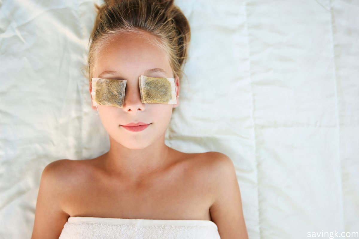 A young woman with fair skin is lying down with used tea bags placed over her eyes as a natural beauty treatment. She is wrapped in a towel and appears relaxed.