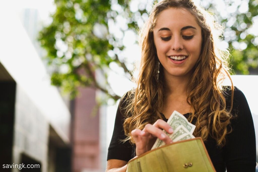 A smiling teenage girl with curly hair pulls dollar bills from her green wallet while standing outdoors.