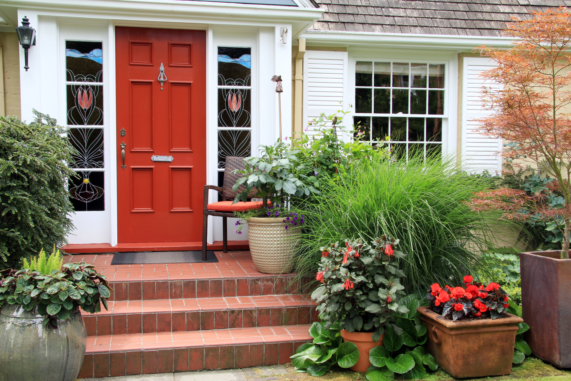 A red front door with elegant stained glass side panels, surrounded by lush greenery and potted plants. In Feng Shui, a red door symbolizes wealth, prosperity, and financial success.