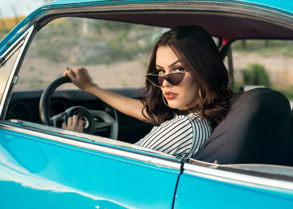 A stylish woman wearing sunglasses sits in the driver's seat of a vintage blue car, looking confidently over her shoulder.