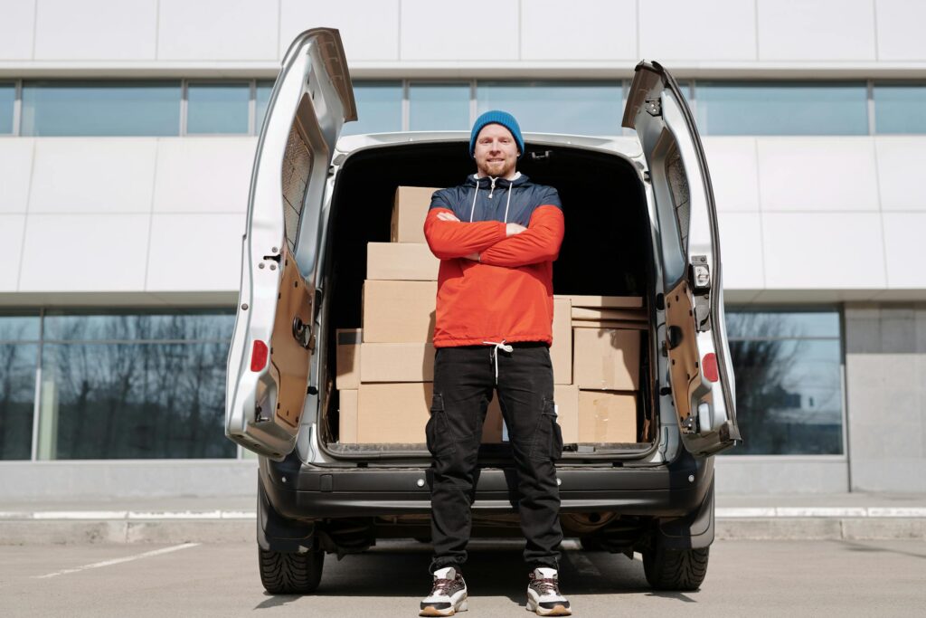 A confident delivery driver standing in front of an open van filled with stacked cardboard boxes, ready for hotshot trucking deliveries.