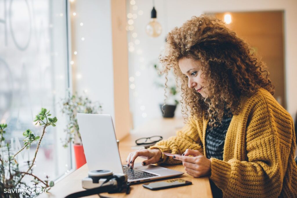 A woman with curly hair wearing a mustard yellow sweater shops online using her laptop while holding a credit card in a cozy café setting.