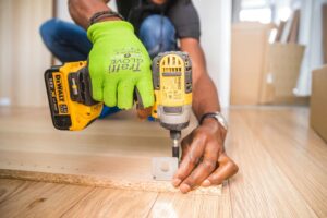 Close-up of a person using a power drill on a wooden board while wearing a green work glove.