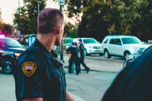 Police officer in uniform standing on a street during a public safety situation, with other officers and vehicles in the background.