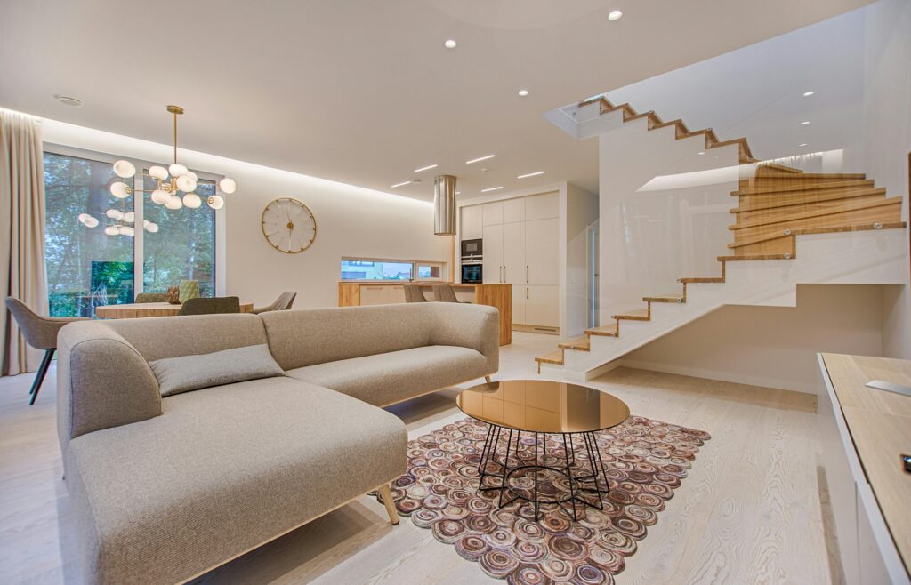 Modern open-concept living space featuring minimalist furniture, floating wood staircase with glass railing, and round gold coffee table on patterned rug.