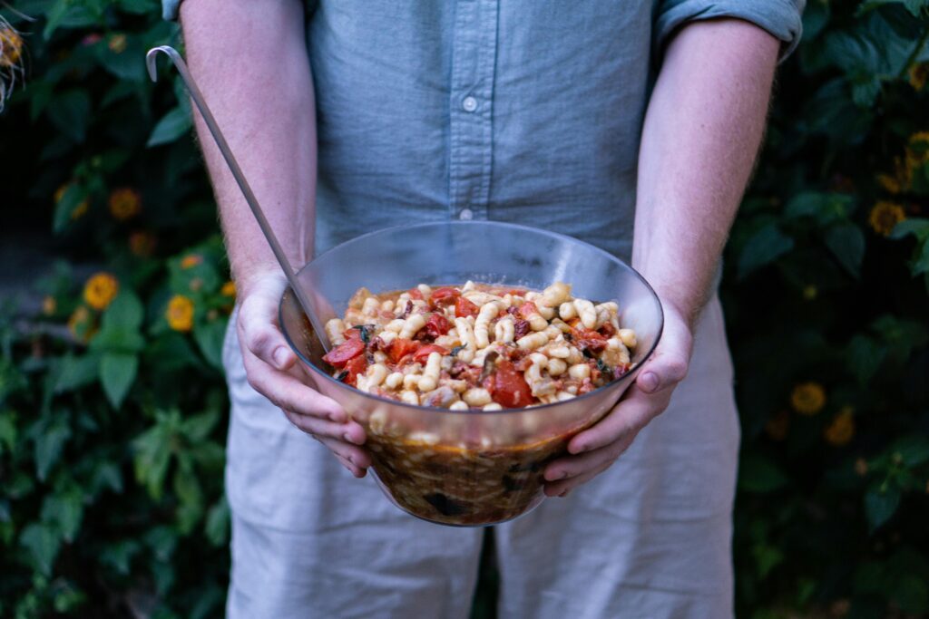 Person holding a large glass bowl of cooked pasta mixed with tomatoes and sauce, with a serving ladle inside.